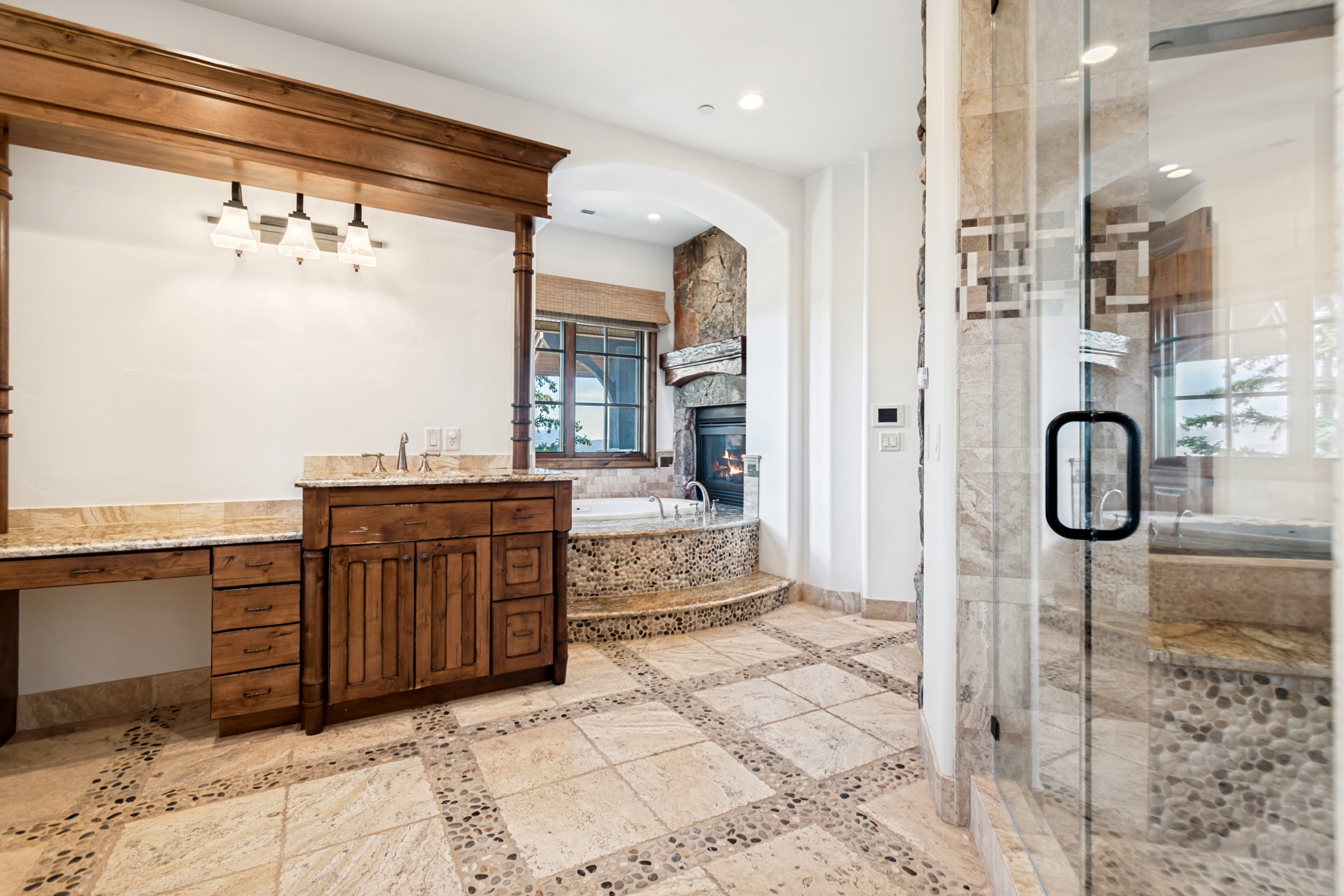 Spacious bathroom featuring stone tile flooring with pebble accents, a deep soaking tub with stone surround, a stone fireplace, wood-finish cabinetry, and a glass-enclosed shower