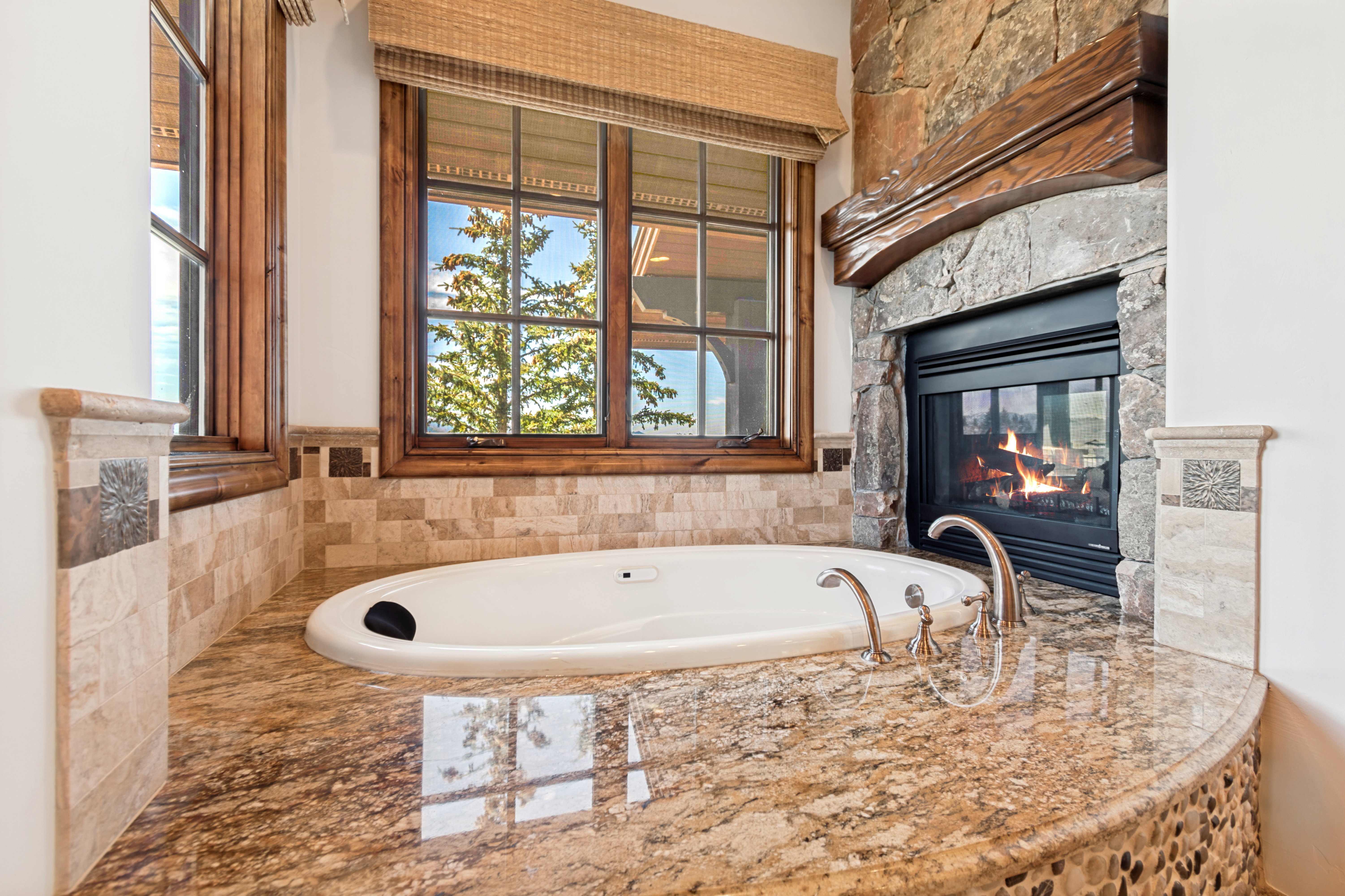 Built-in soaking tub with deck-mounted fixtures, surrounded by a stone-faced fireplace with a substantial wood mantel, a polished stone countertop, and natural wood-framed windows