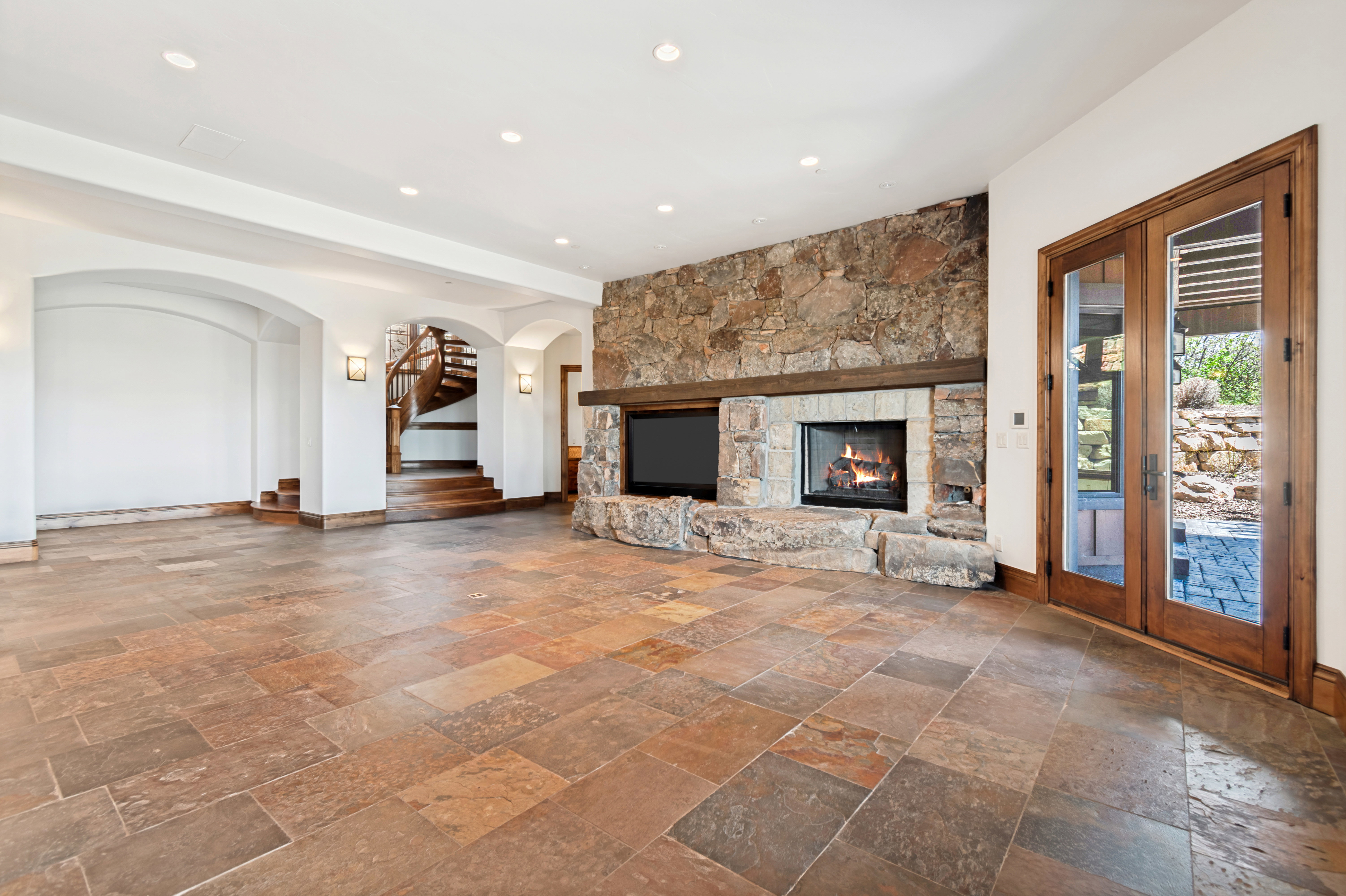 Spacious room featuring large format natural stone tile flooring, a prominent stone fireplace with wood mantel, recessed lighting, and wooden double doors