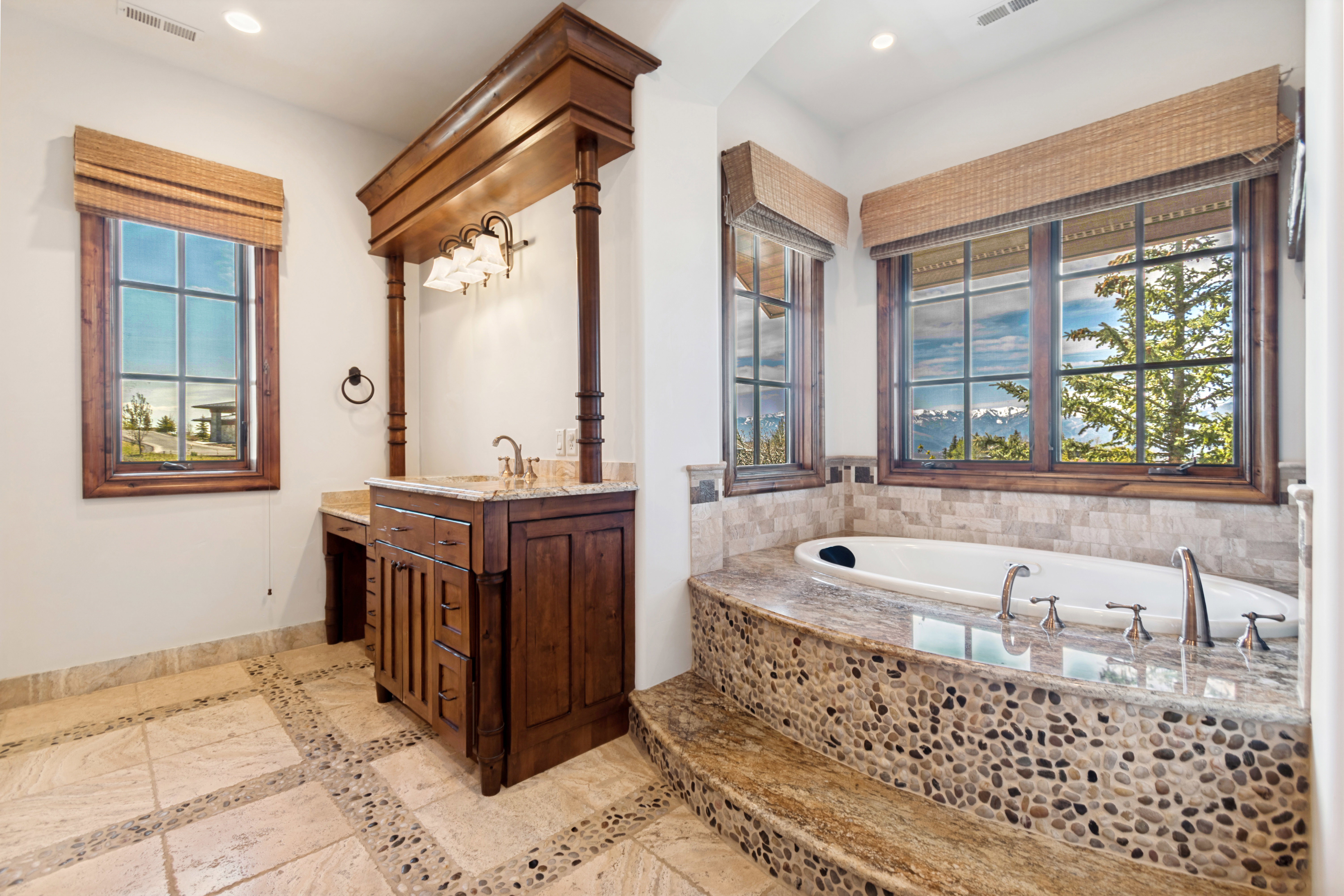 Bathroom featuring an integrated soaking tub with stone surround, a wood vanity with a stone countertop, a separate vanity area, and two large windows with natural wood frames