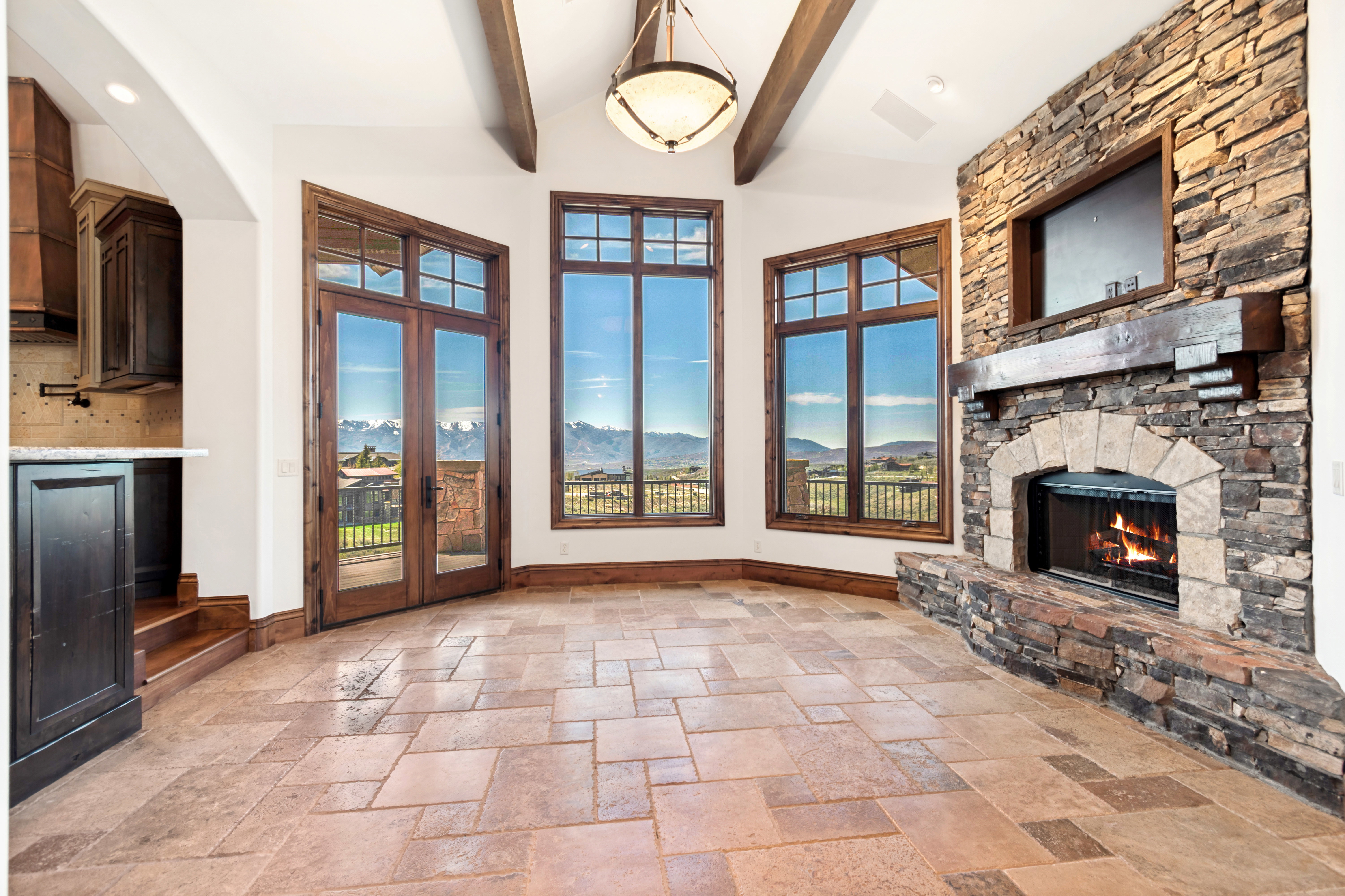 Spacious living area featuring a stone-clad fireplace with a timber mantel, natural light from multiple large windows, a glass-paneled double door, a vaulted ceiling with exposed wood beams, and patterned tile flooring