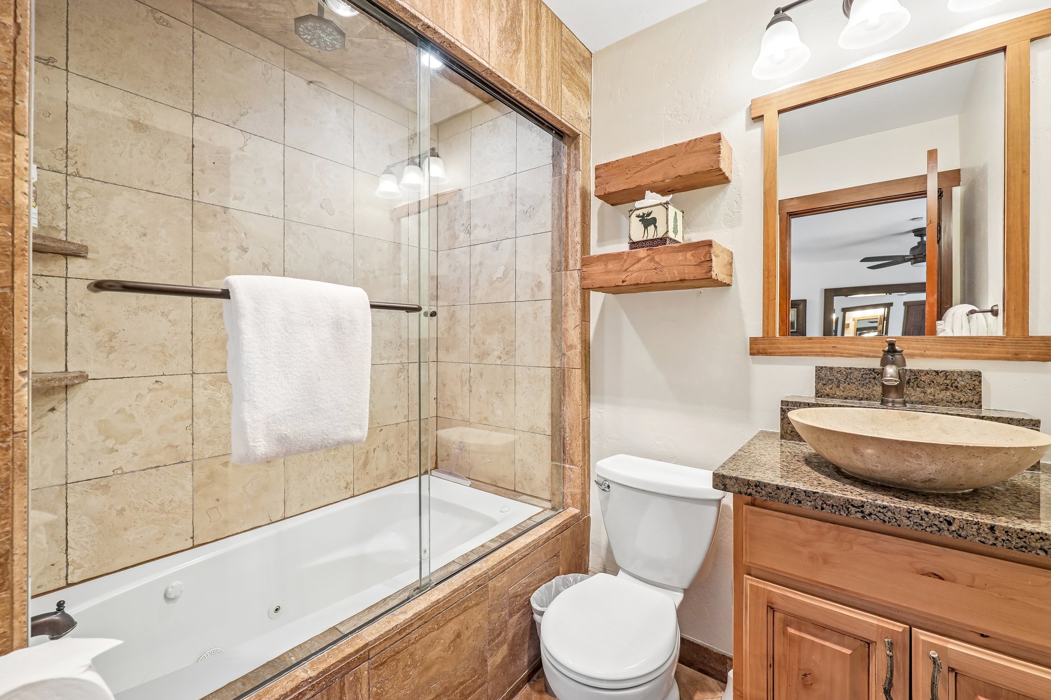 Bathroom featuring a jetted tub with glass enclosure, stone-tiled shower surround, a vessel sink on a granite-look countertop, and a wood-framed mirror