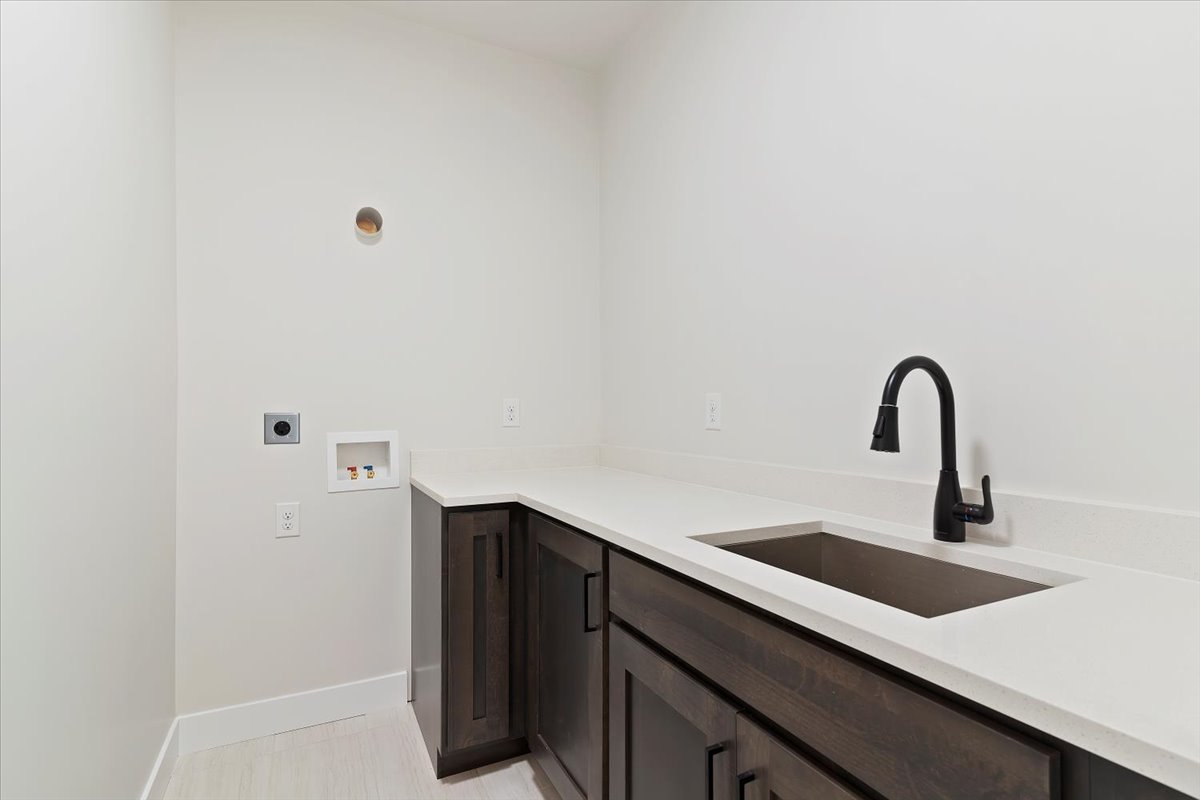 Laundry room featuring dark wood-finish cabinetry, light-toned countertops, an undermount sink with a matte black faucet, and light wood-finish flooring