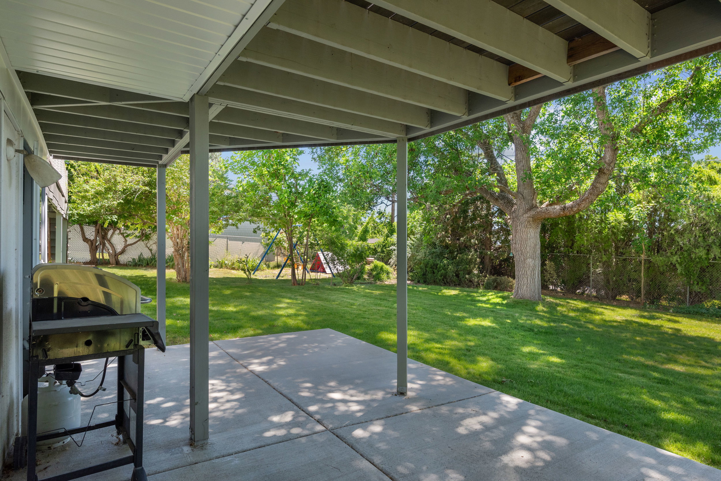 Covered concrete patio overlooking a spacious lawn with mature trees and perimeter fencing