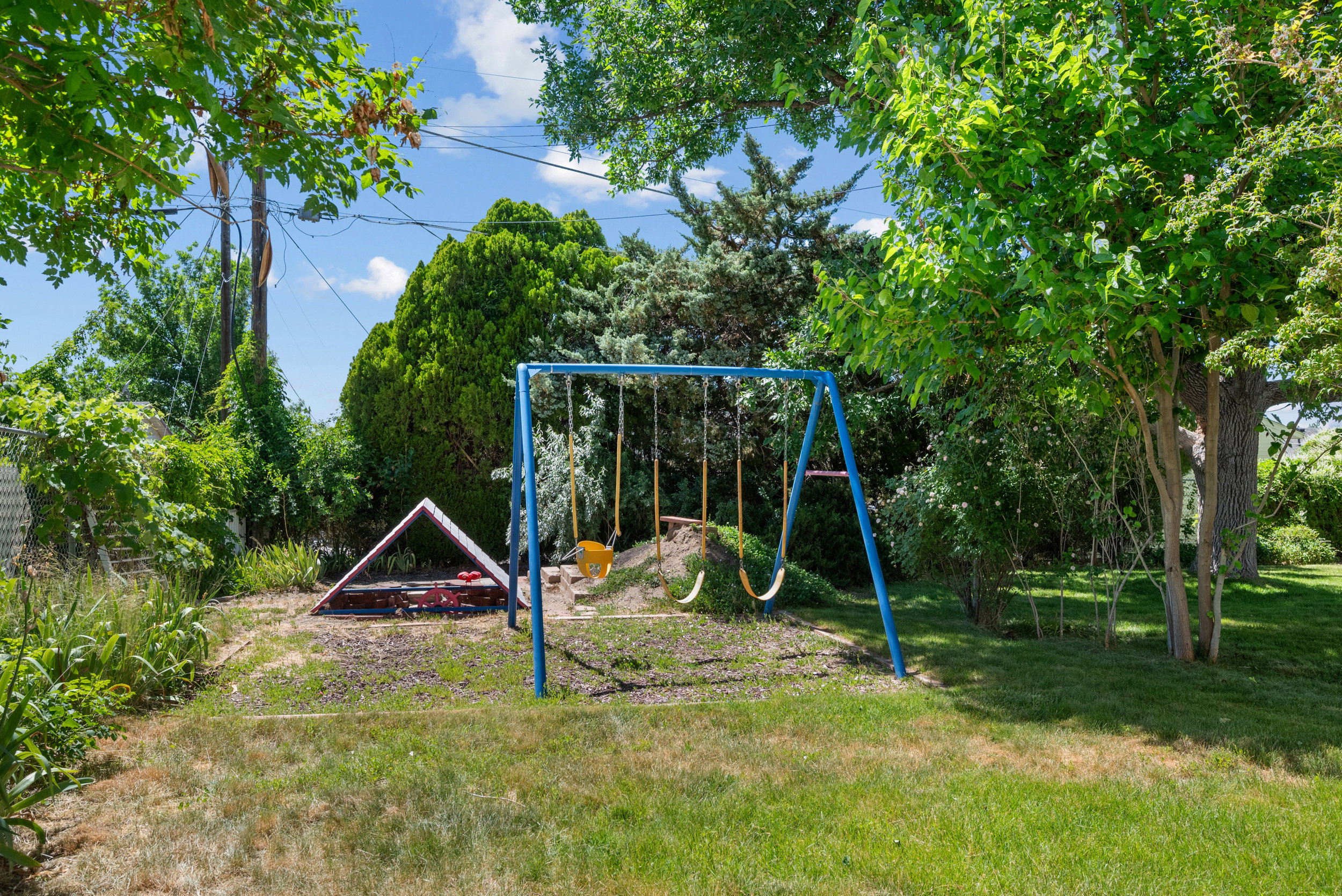 Spacious backyard featuring a blue swing set, a red sandbox with an A-frame cover, and mature green trees providing natural shade