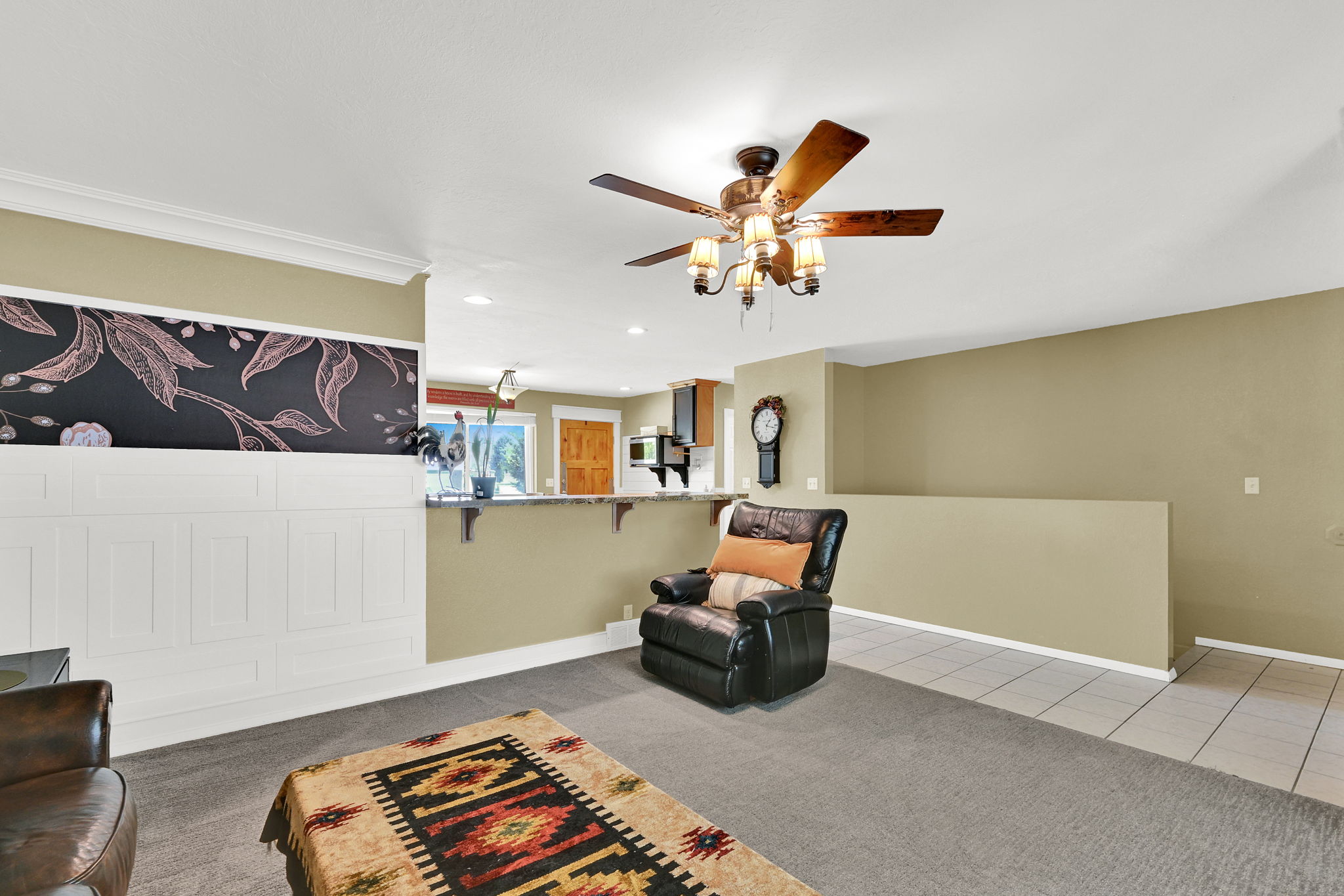 Open concept living area featuring a ceiling fan with integrated lighting, a decorative half-wall with paneling and a botanical-themed accent, and a breakfast bar with a stone-finish countertop