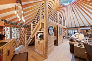 Spacious circular interior featuring exposed timber ceiling beams, natural wood flooring, a built-in wooden staircase with black metal railings, a skylight, and integrated storage cabinetry