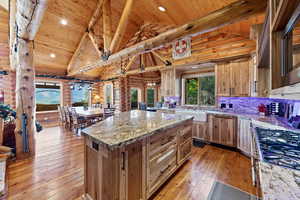 Open-concept kitchen and dining area featuring natural wood walls, vaulted ceilings with exposed beams, wood-finish flooring, a central island with a stone countertop, and a farmhouse sink