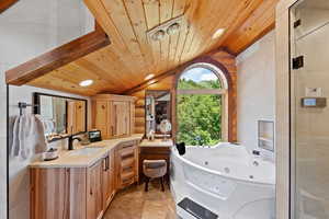 Spacious bath featuring an arched window, built-in jetted tub, wood-finish ceiling, exposed wood beams, and a double vanity with stone countertops