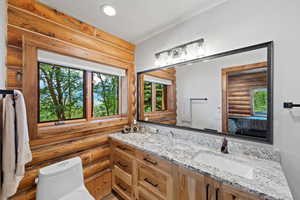 Bathroom featuring log wall construction, a large picture window, a double vanity with a granite countertop, and a wide framed mirror with integrated lighting