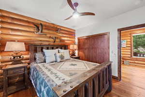 Bedroom featuring log wall construction, wood-finish flooring, a wood-paneled closet with sliding doors, an open doorway with wood trim, and a ceiling fan with integrated lighting