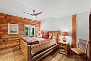 Bedroom featuring log wall construction, wood-finish flooring, a window with exterior views, a ceiling fan with integrated lighting, and a prominent wood beam detail