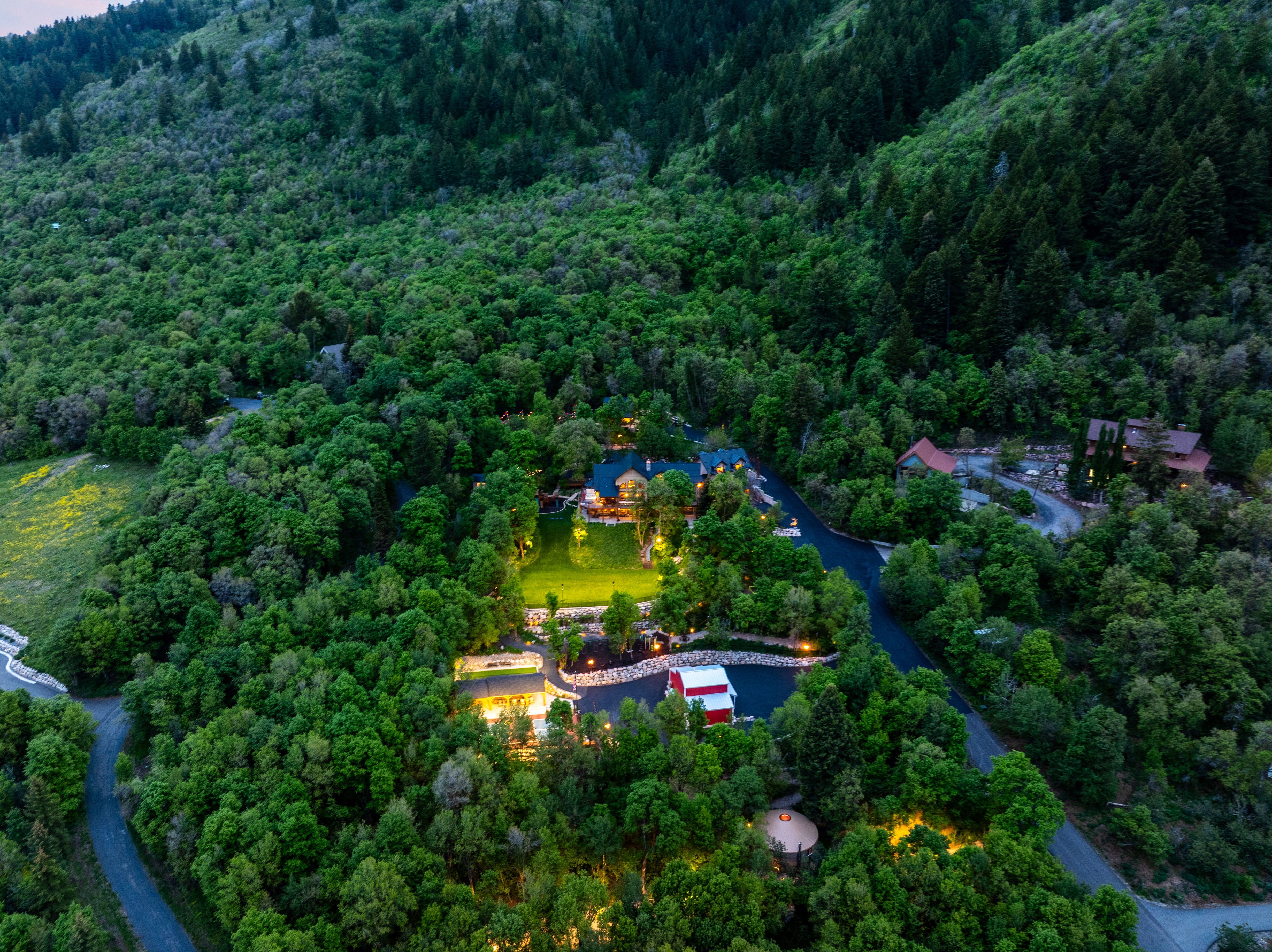 Expansive property nestled among mature trees, featuring multiple structures, paved driveways, a central illuminated lawn, and surrounding forested hills