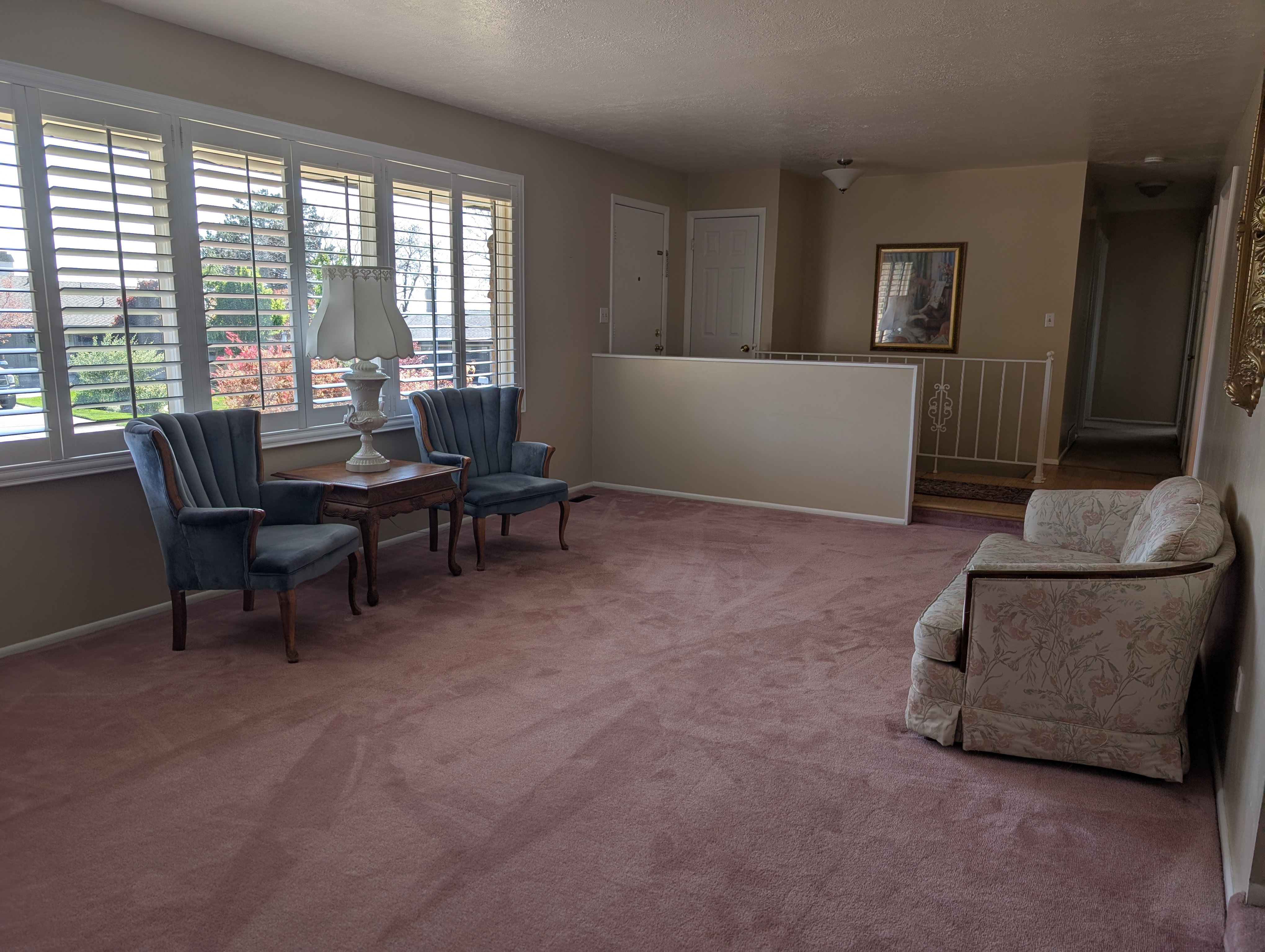 Spacious living room featuring a wall of shuttered windows, plush carpeting, and a built-in half-wall with a decorative metal railing