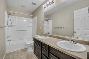 Bathroom featuring a dual vanity with integrated sinks, a tub/shower combination with built-in shelving, dark wood-finish cabinetry, and tiled flooring