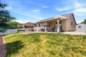 Rear exterior featuring a covered patio with black railings, a gravel and rock fire pit area, a white vinyl fence, and a detached shed on RV pad.