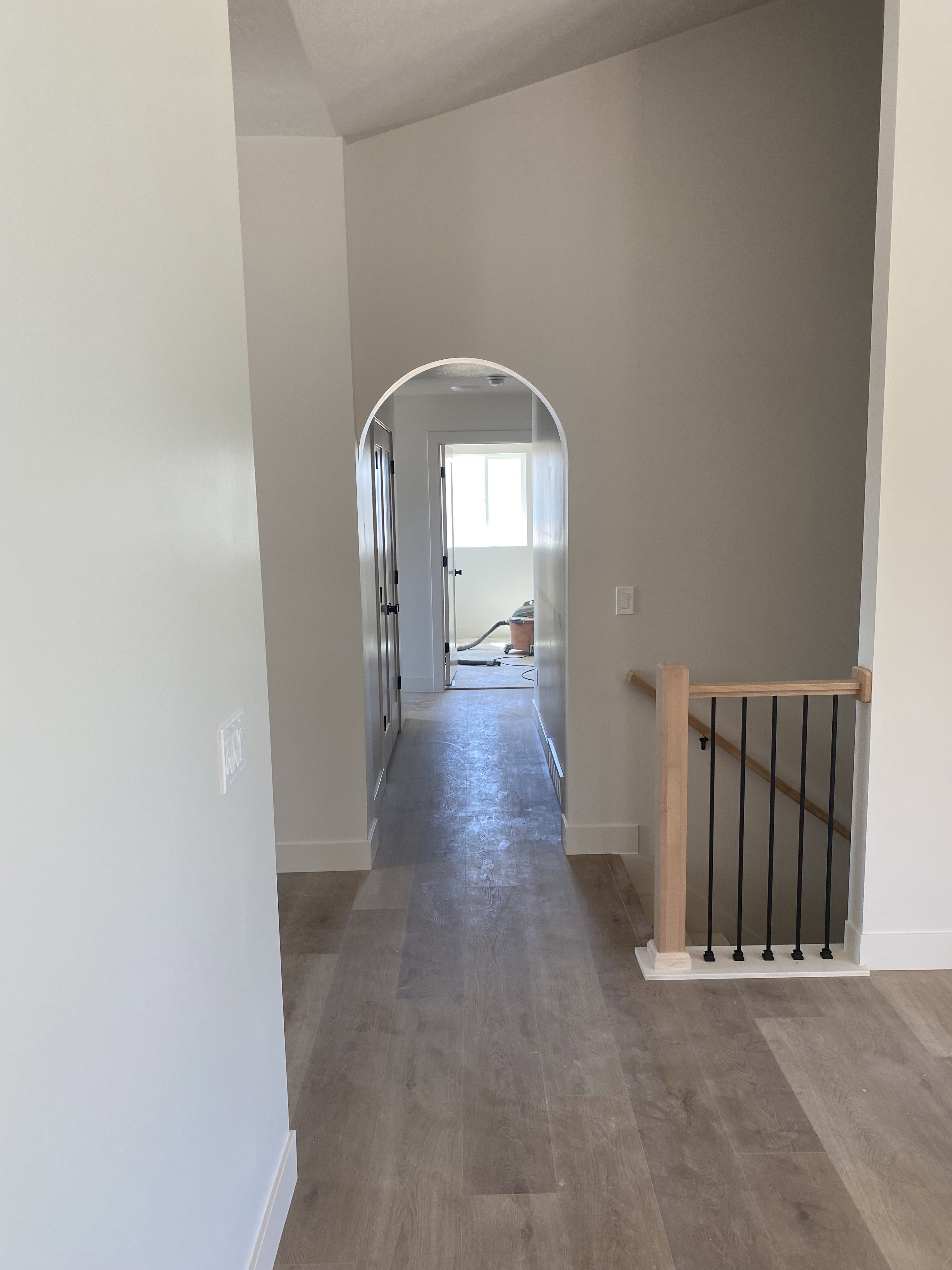 Arched doorway opening to a hallway featuring wood-finish flooring, crisp white baseboards, and a natural wood banister with black iron balusters