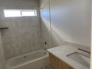 Bathroom featuring a built-in bathtub with subway tile surround, integrated window, light wood-finish vanity with gold-tone hardware, and a stone countertop with undermount sink