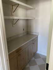 Pantry featuring floating white shelving with brass brackets, light wood-finish cabinetry, light-toned countertop, and checkered tile flooring