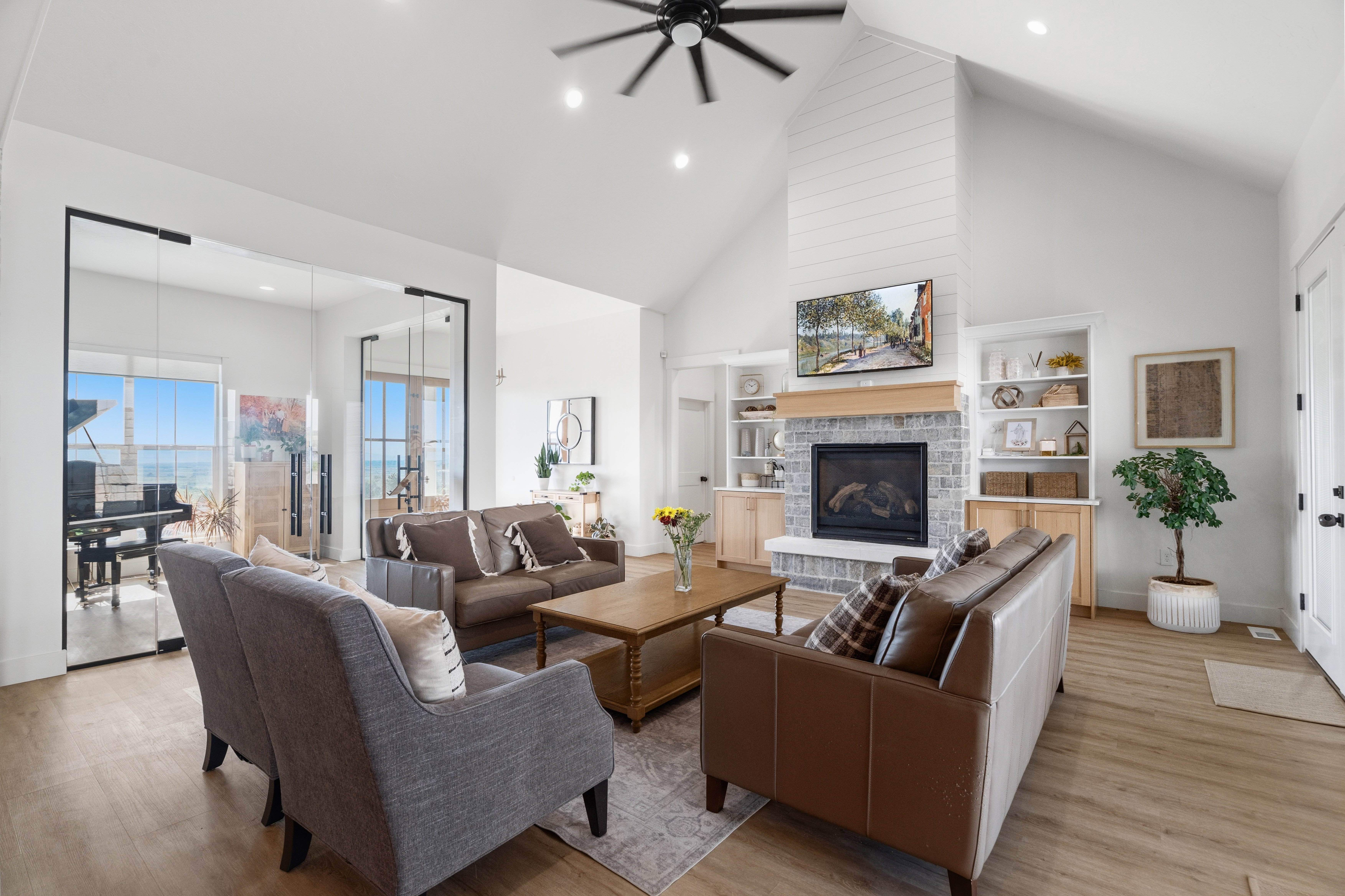 Vaulted ceiling with a black ceiling fan, fireplace with a shiplap chimney and stone surround, wood-finish flooring, built-in shelving, and glass French doors leading to a separate room