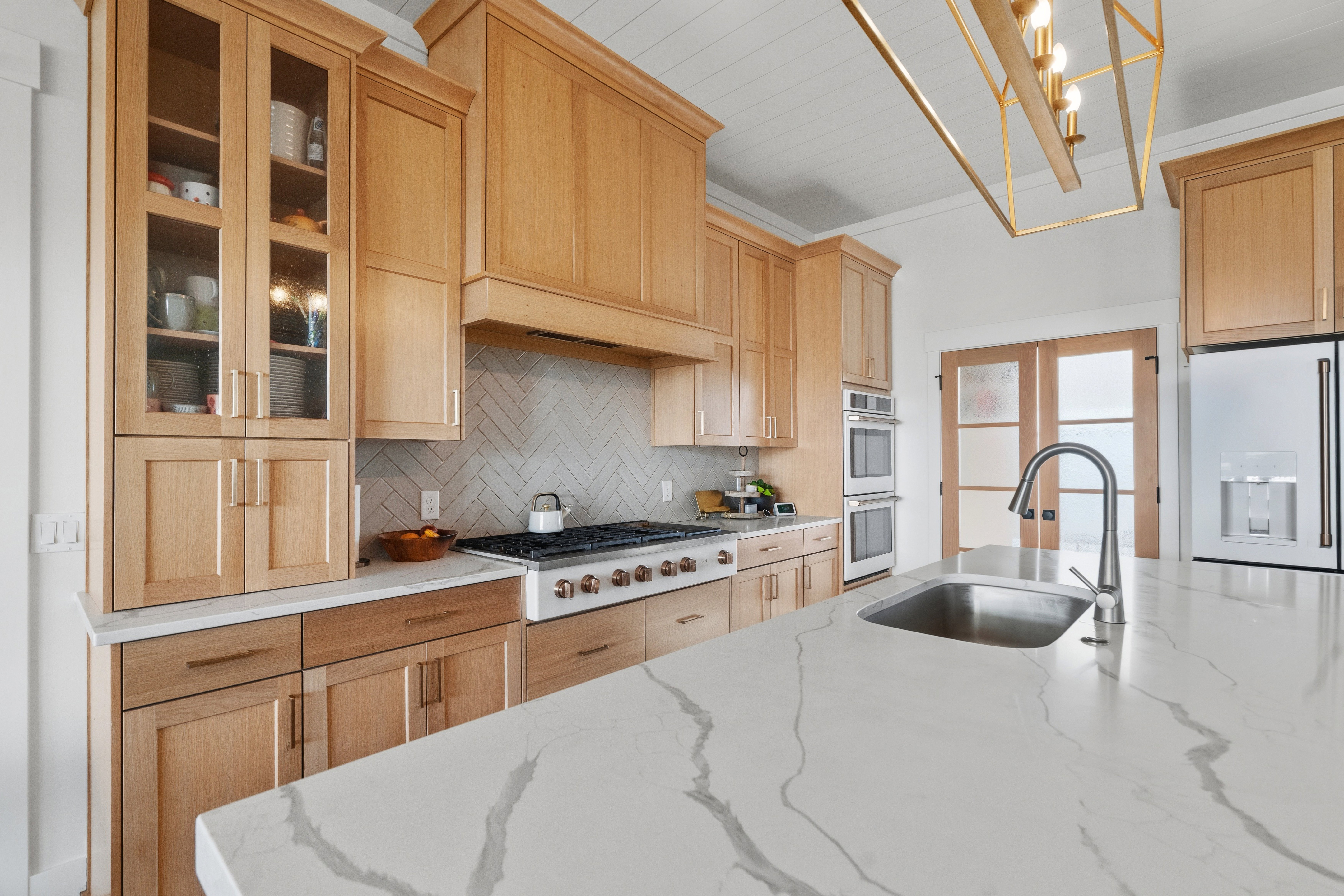 Spacious kitchen featuring a large island with an undermount sink, light wood cabinetry, white countertops, built-in double ovens, and a gas range with a herringbone tile backsplash