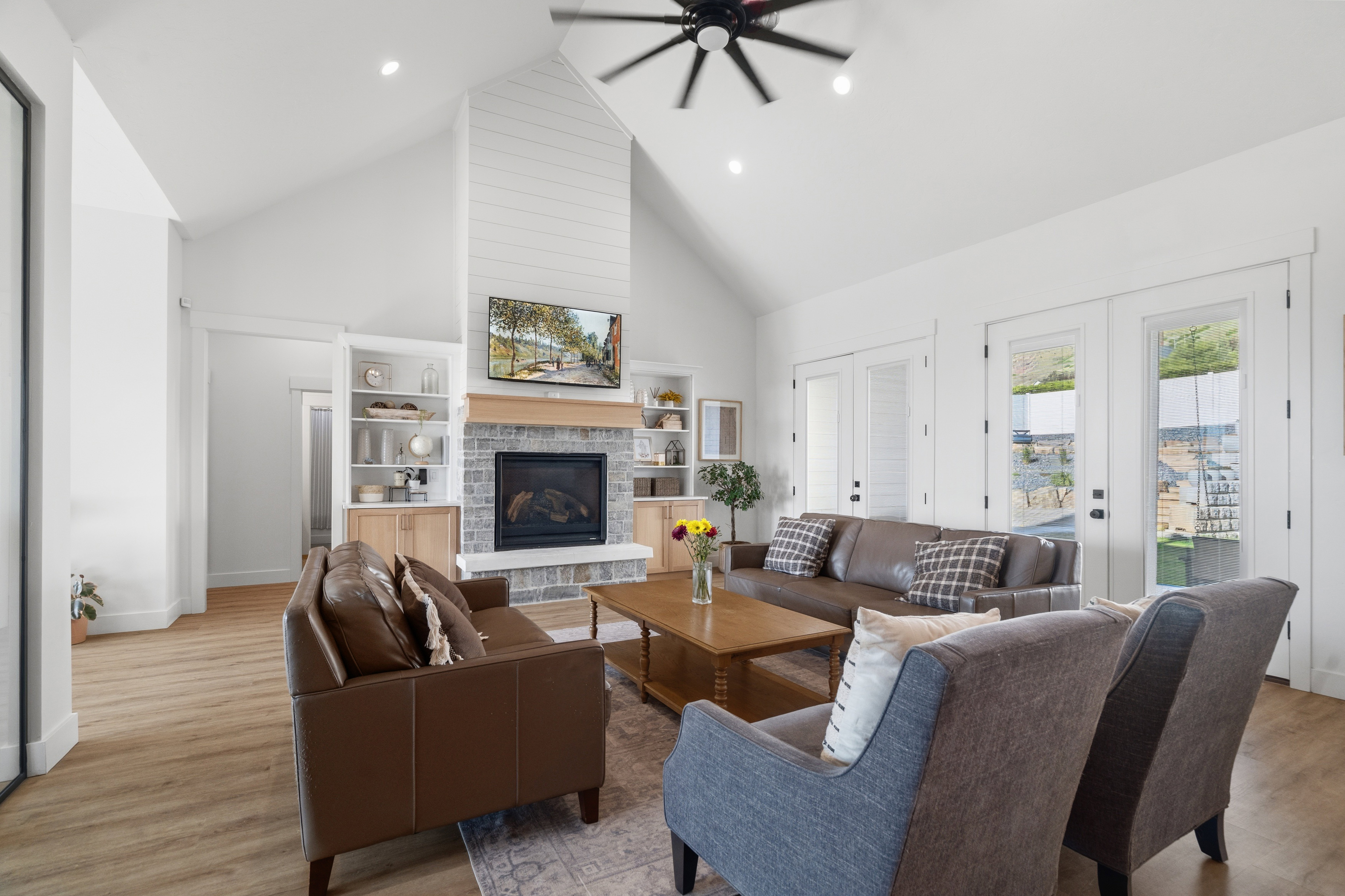 Vaulted ceiling living area featuring a stone and shiplap fireplace, wood-finish flooring, built-in shelving, and multiple glass-paneled doors