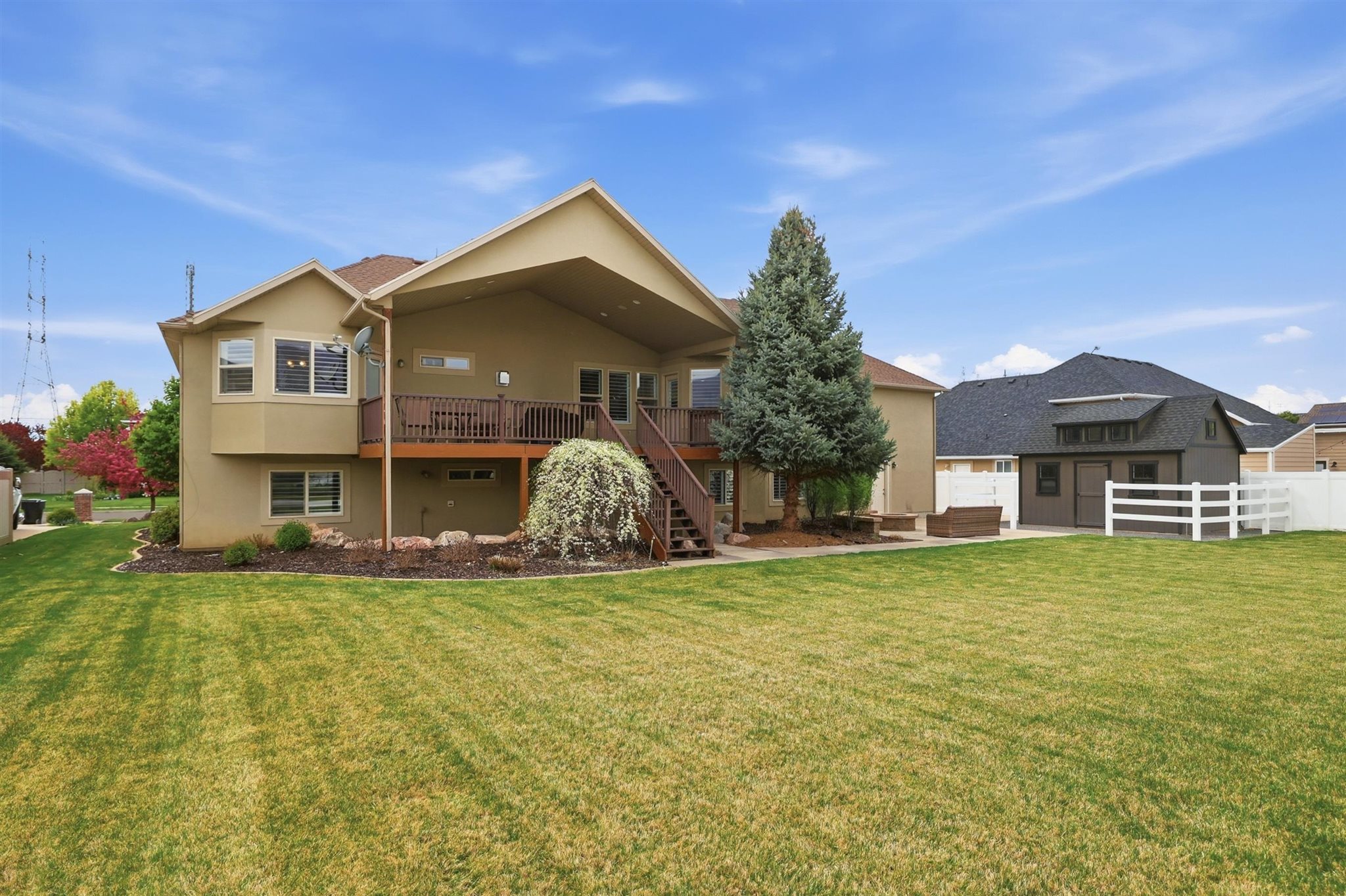 Spacious backyard featuring a large green lawn, a multi-level deck with stairs, mature landscaping, and a detached shed with a white picket fence
