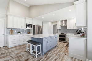 Modern kitchen featuring a blue island with a white countertop, white cabinetry with silver hardware, stainless steel appliances, wood-finish flooring, and a vaulted ceiling