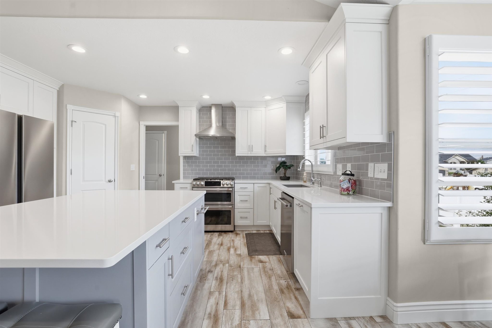 Contemporary kitchen featuring a large central island with a white countertop, white shaker-style cabinetry, stainless steel appliances, a gray subway tile backsplash, and wood-finish flooring