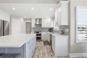 Contemporary kitchen featuring a large central island with a white countertop, white shaker-style cabinetry, stainless steel appliances, a gray subway tile backsplash, and wood-finish flooring