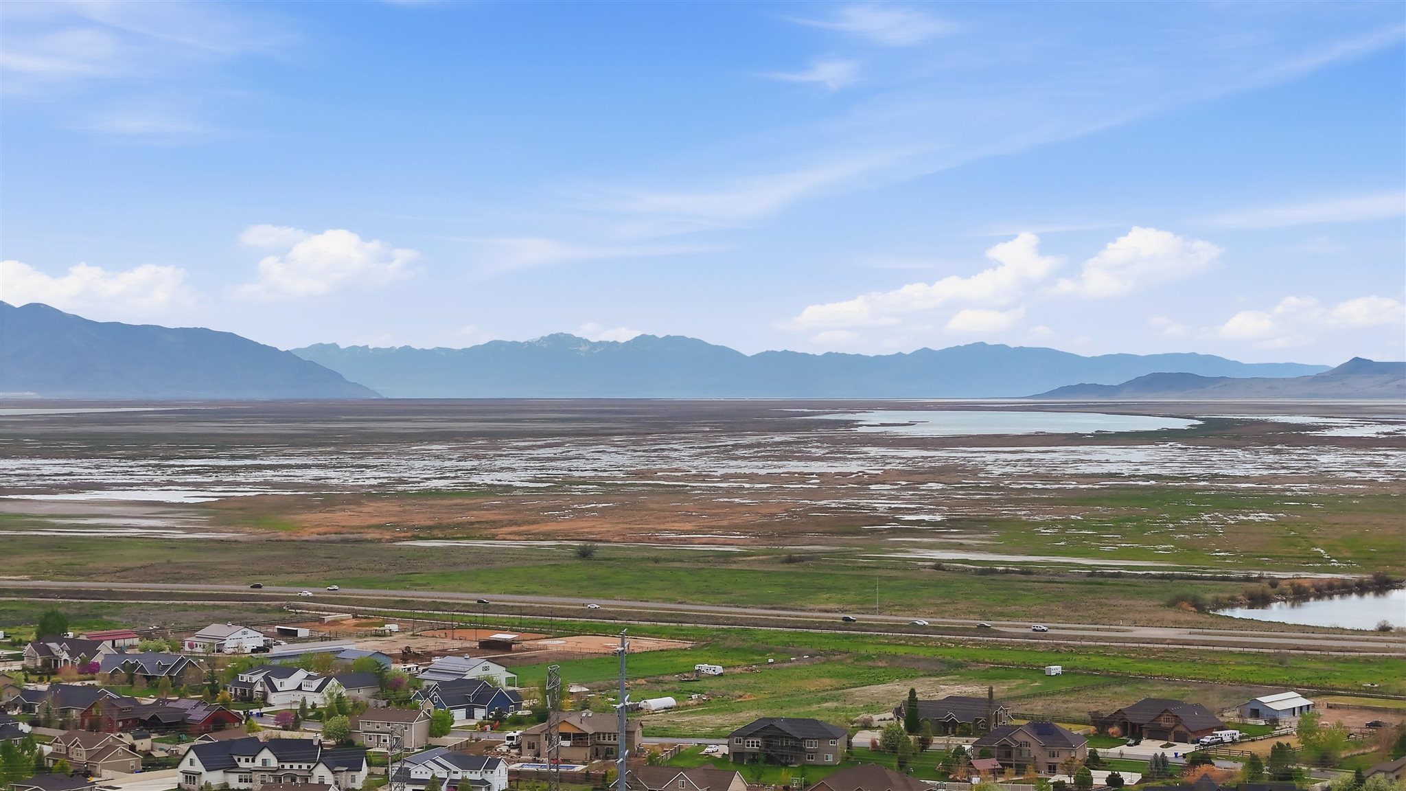 Expansive aerial perspective showcasing a residential neighborhood with diverse architectural styles, framed by distant mountains, a large body of water, and surrounding green landscapes