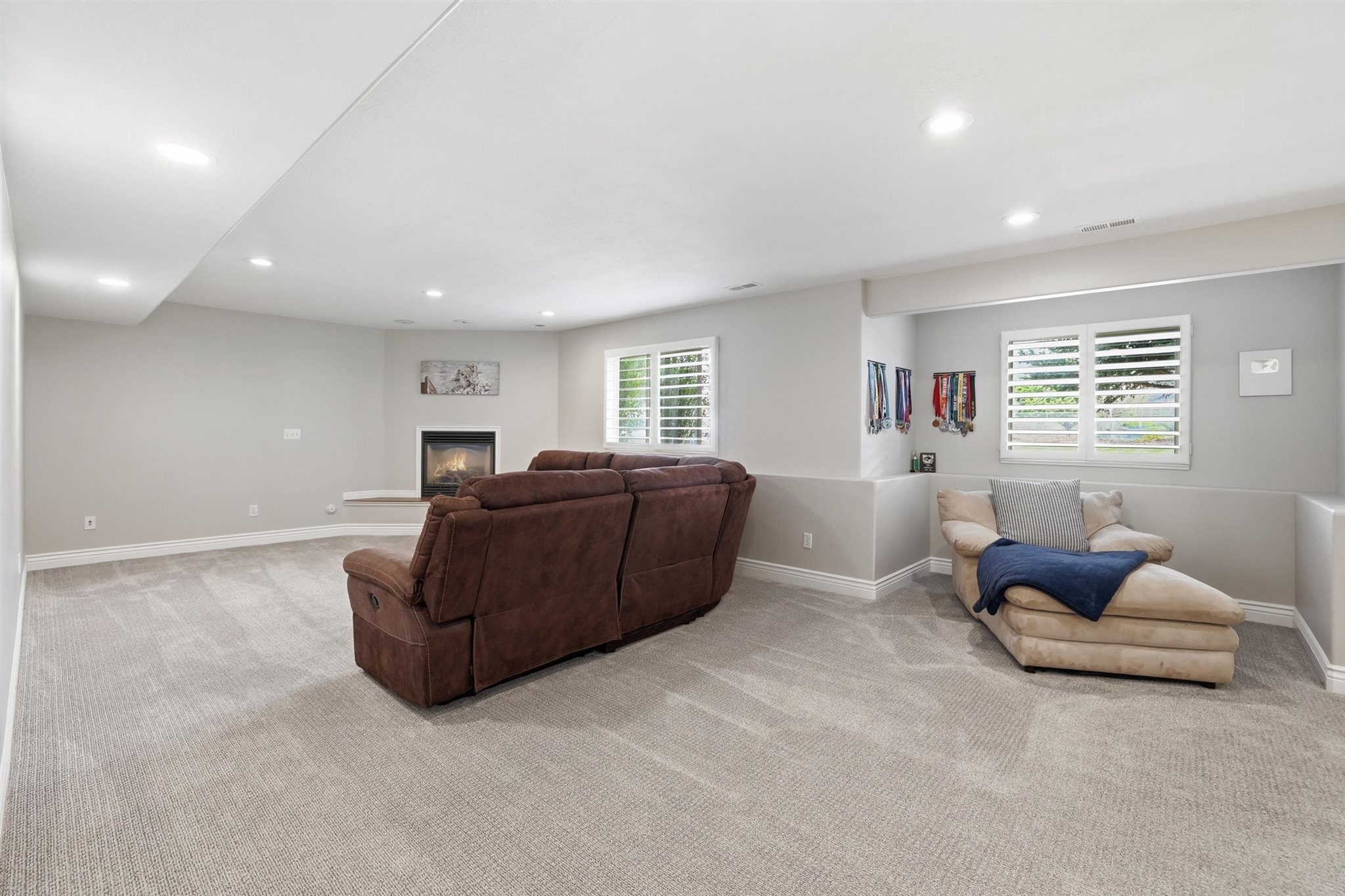 Spacious room featuring light gray carpeting, recessed ceiling lighting, a corner fireplace with a white hearth, and multiple windows with white plantation shutters