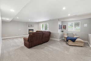 Spacious room featuring light gray carpeting, recessed ceiling lighting, a corner fireplace with a white hearth, and multiple windows with white plantation shutters