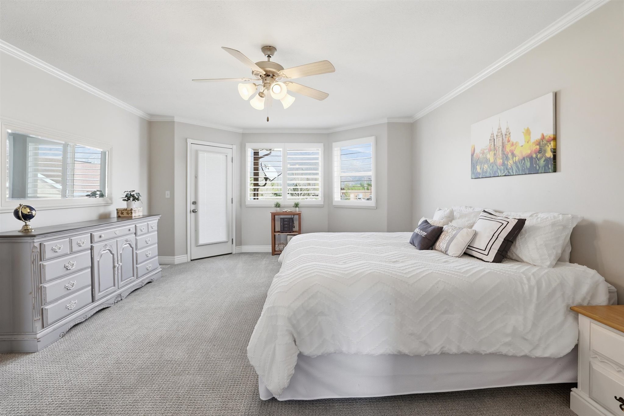 Carpeted bedroom featuring a ceiling fan with integrated lighting, crown molding, bay windows with plantation shutters, and a glass-paneled door