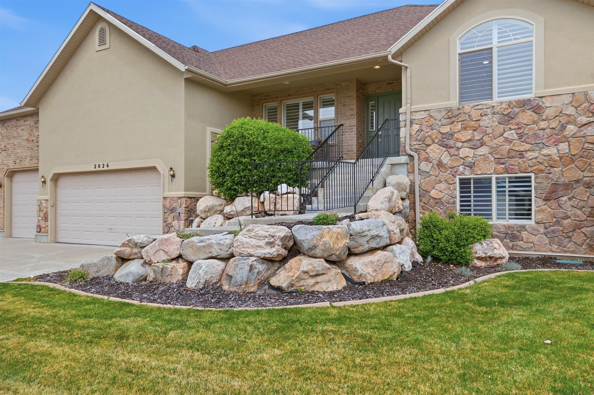 Stucco and stone exterior featuring a two-car garage, arched window, and rock retaining wall with mature landscaping