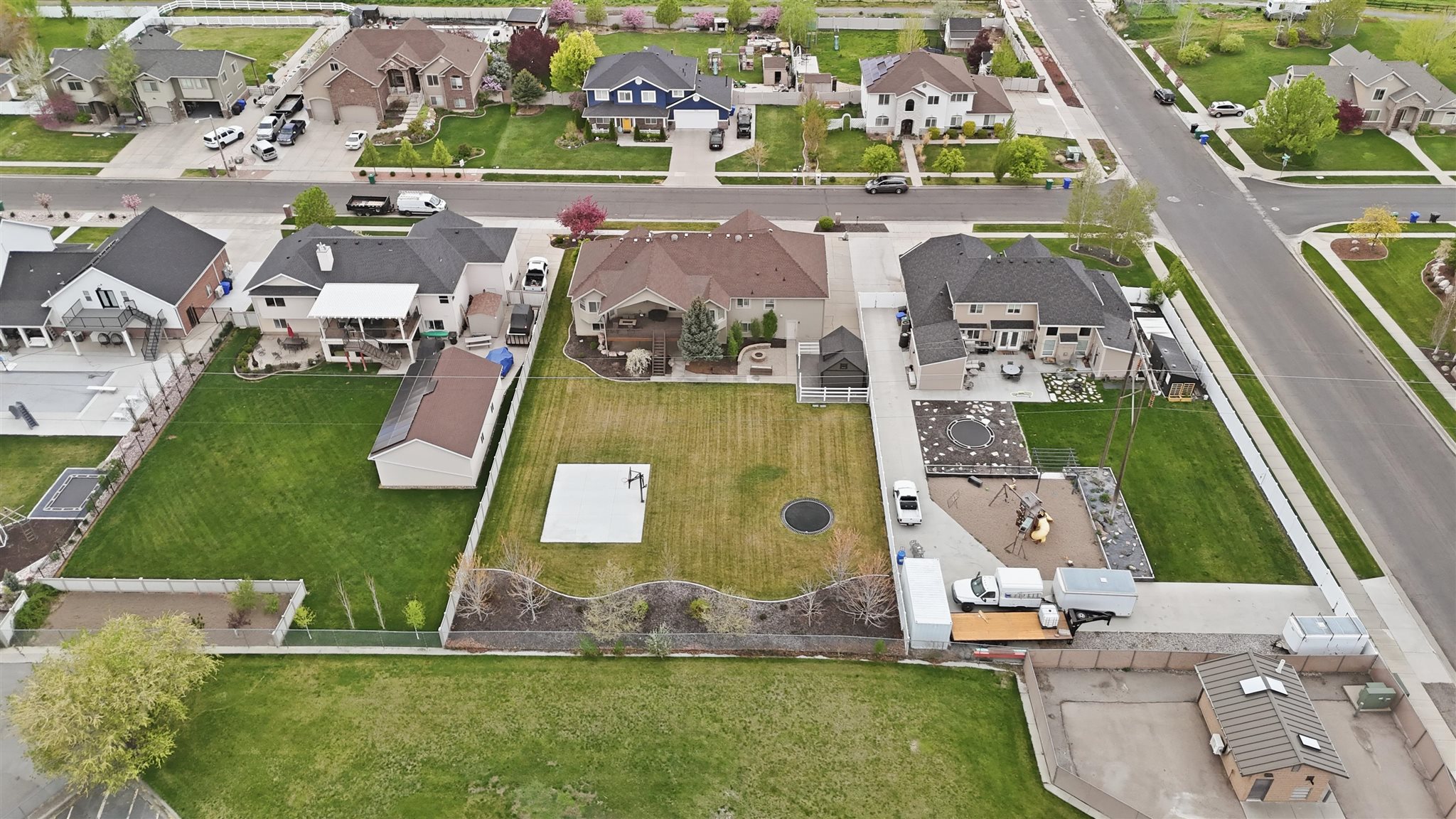 Expansive backyard featuring a large green lawn, distinct patio space, a dedicated play area with equipment, a trampolene, and mature perimeter landscaping