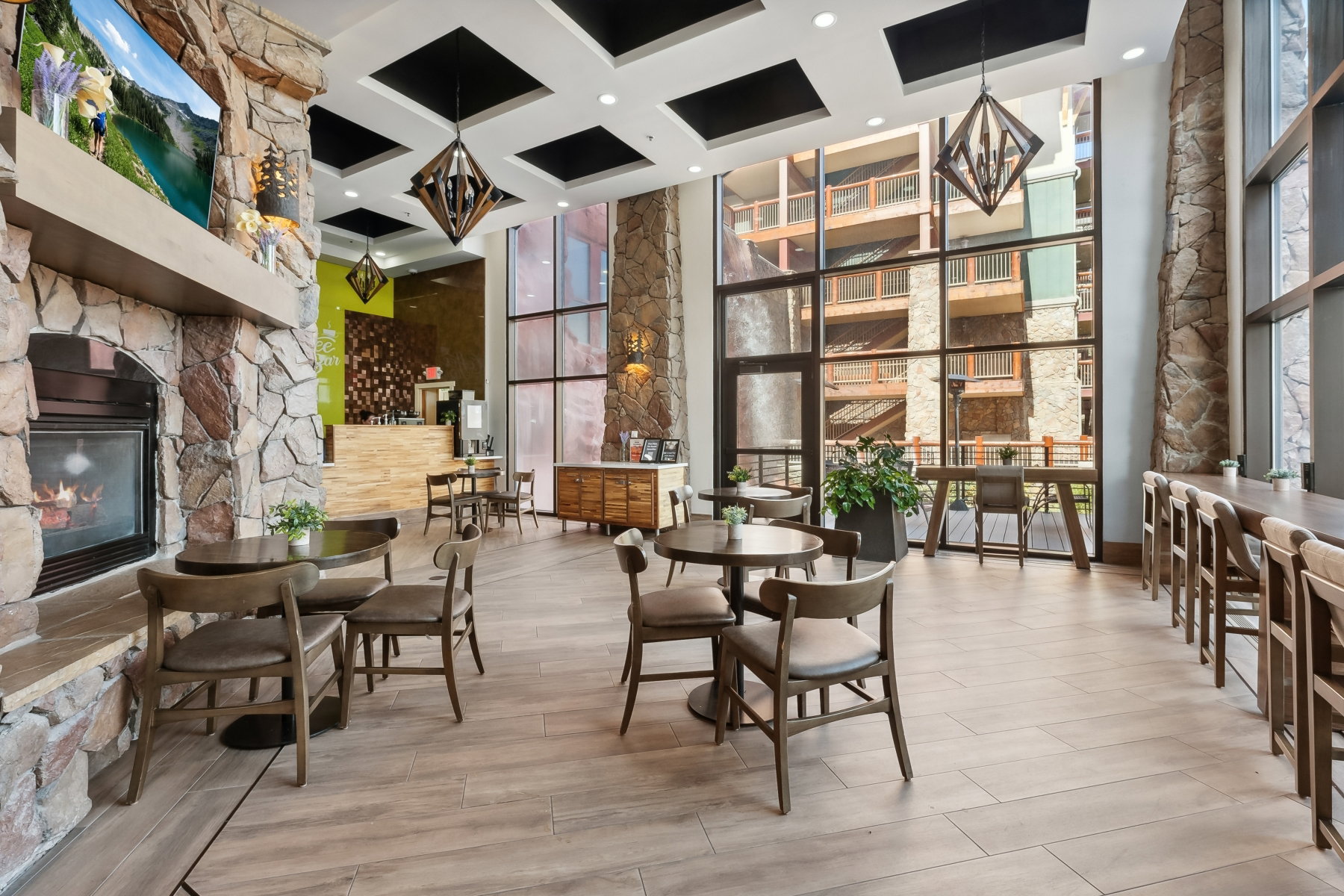 Open-concept common area featuring a stone fireplace, wood-finish flooring, full-height windows, a built-in bar, and coffered ceilings with geometric pendant lighting
