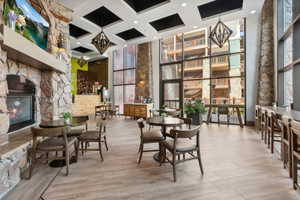 Open-concept common area featuring a stone fireplace, wood-finish flooring, full-height windows, a built-in bar, and coffered ceilings with geometric pendant lighting