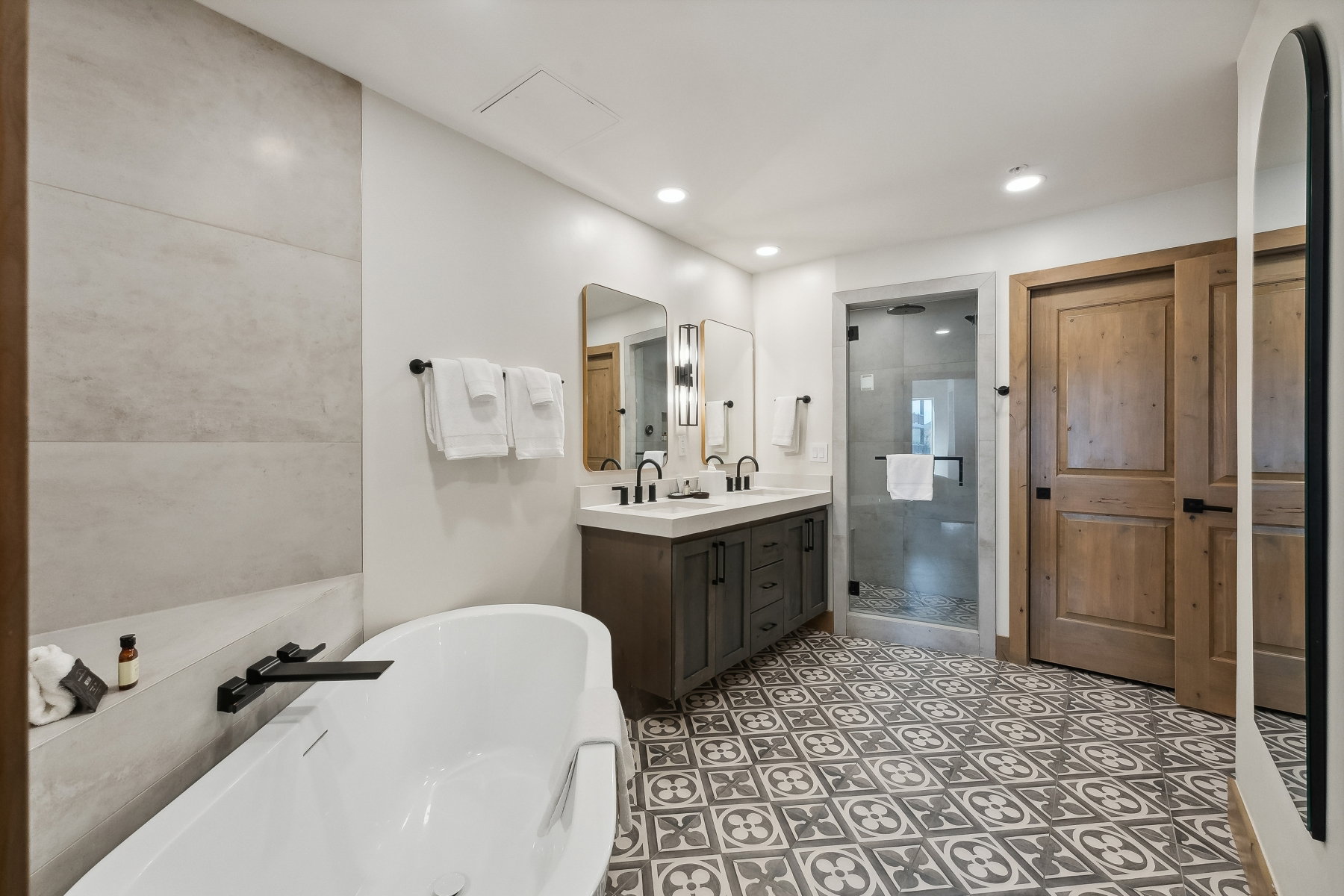 Contemporary bathroom featuring a freestanding soaking tub with a matte black faucet, a dual vanity with a white countertop, a walk-in shower with a glass enclosure, and patterned floor tiling