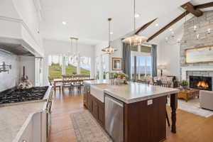 Open-concept kitchen and living area featuring high vaulted ceilings with exposed wood beams, a stone-clad fireplace with a white mantel, and engineered hardwood flooring