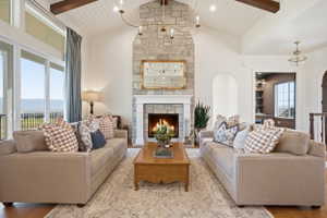 Vaulted ceiling with exposed wood beams, stone fireplace with a white mantel, sliding glass doors, and engineered hardwood flooring