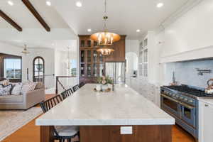 Spacious kitchen island with a marble countertop, a professional-grade range, full-height white cabinetry with glass inserts, and wood-finish ceiling beams