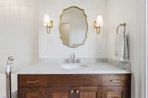 Bathroom vanity featuring a wood-finish cabinet with brass hardware, a white countertop, an undermount sink with a polished nickel faucet, and a decorative mirror flanked by two sconces
