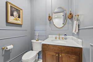 Powder room featuring panel molding, a wood-finish vanity with a stone countertop, a gold-framed mirror, and brass hardware