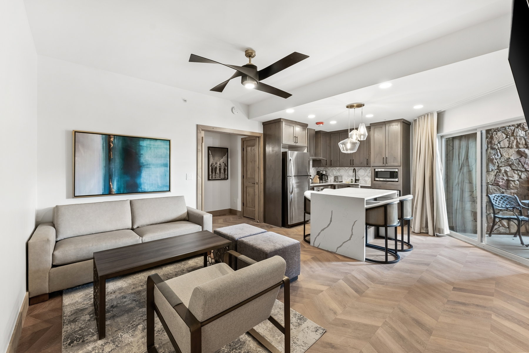 Open-concept living space featuring herringbone wood-finish flooring, a modern kitchen island with waterfall countertop, stainless steel appliances, and sliding glass doors opening to a private patio with stone wall