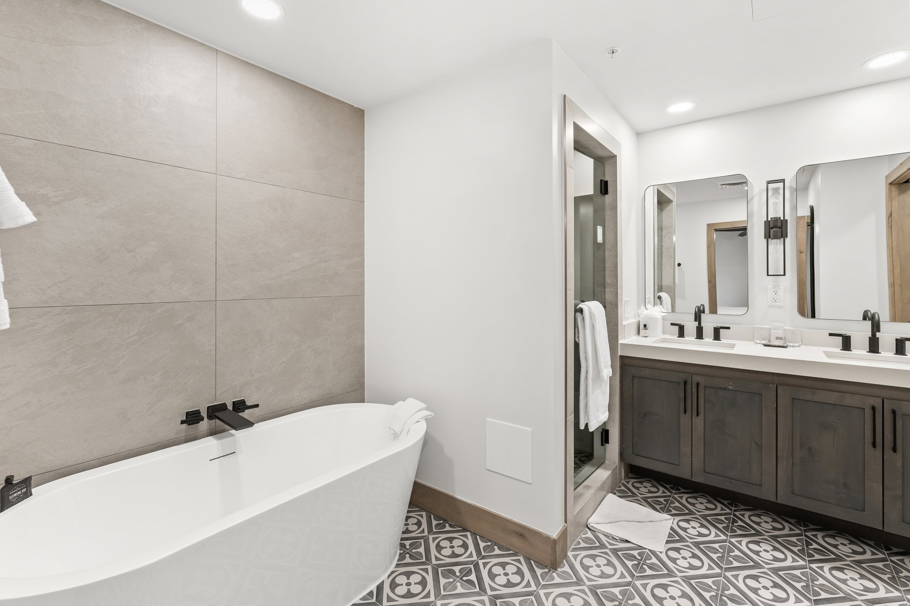 Contemporary bathroom featuring a freestanding soaking tub with a wall-mounted matte black faucet, an expansive vanity with double sinks, and patterned floor tiles