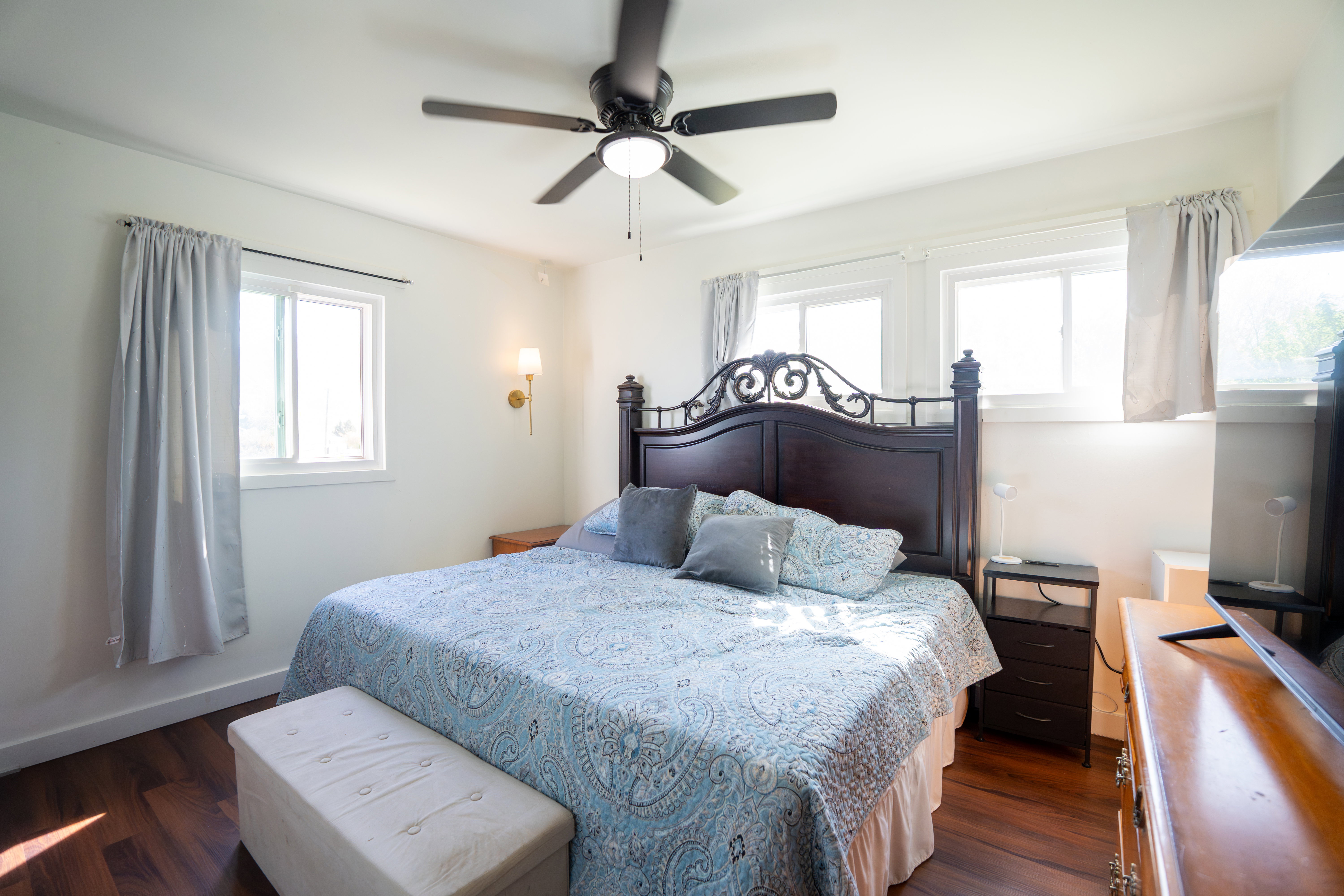 Charming bedroom featuring dark wood-finish flooring, white walls, and bright windows allowing for natural light