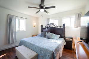 Charming bedroom featuring dark wood-finish flooring, white walls, and bright windows allowing for natural light