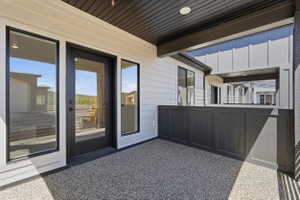 Covered patio featuring a textured aggregate surface, dark paneled privacy wall, white horizontal siding, recessed lighting, and black-framed glass door