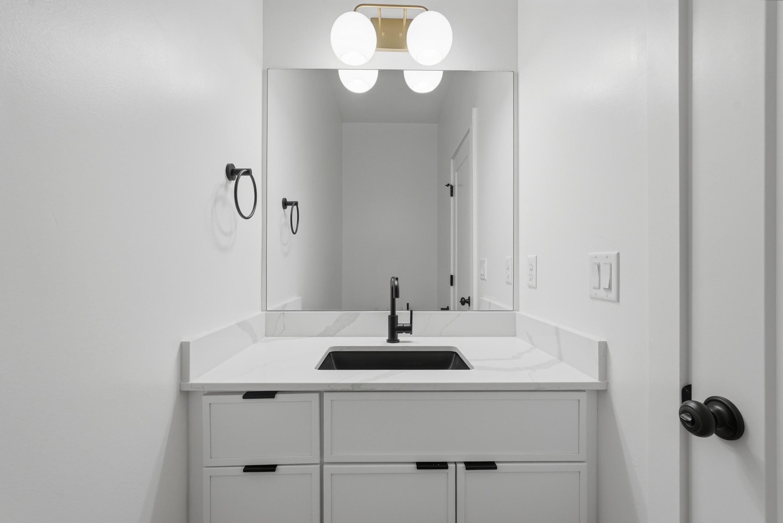 Modern vanity with a sleek black sink, white countertop, white cabinetry with black hardware, and an overhead gold-finish light fixture